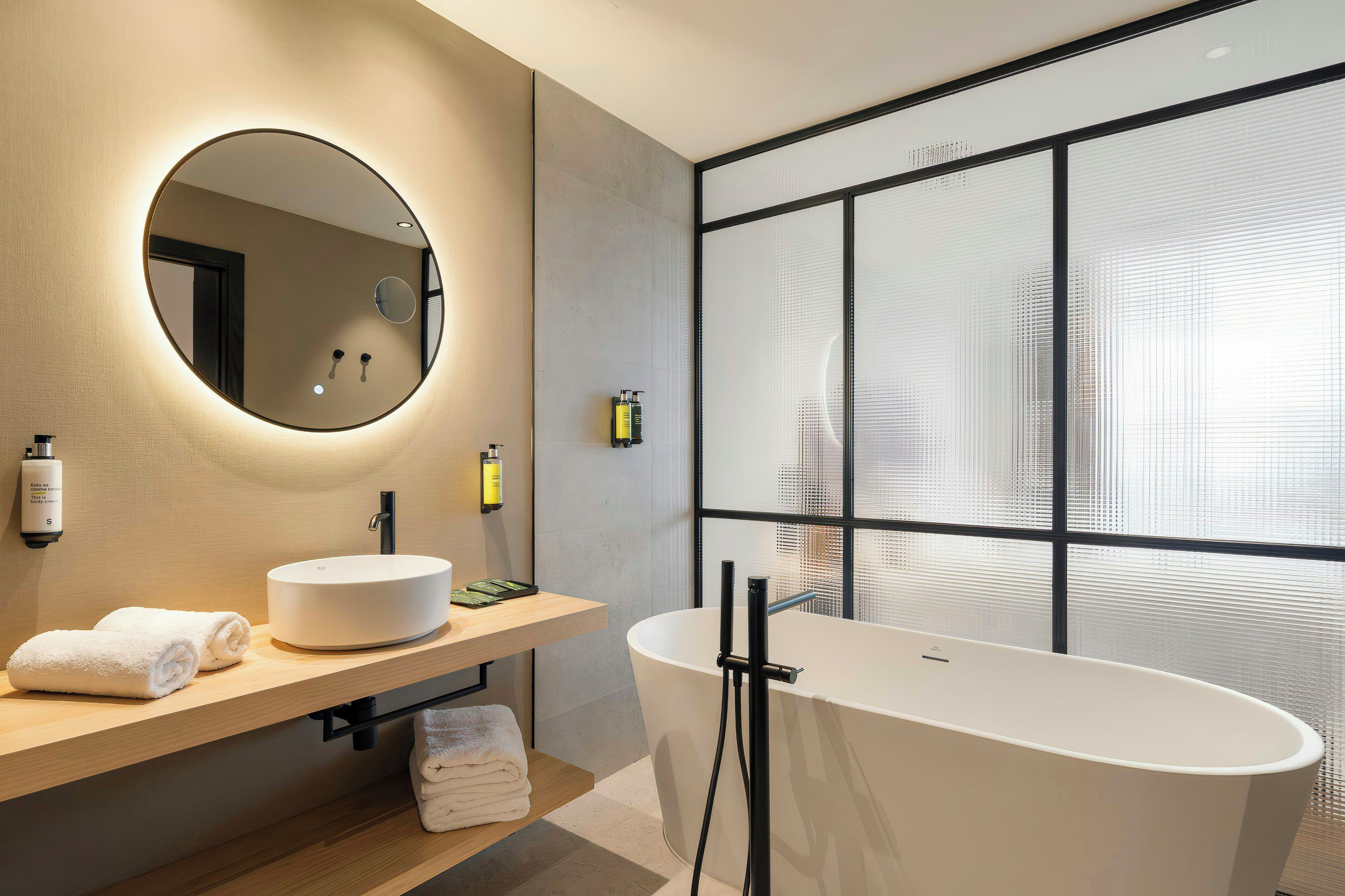 Een moderne badkamer met een ronde spiegel, een vrijstaand bad en een groot raam met zonwering.