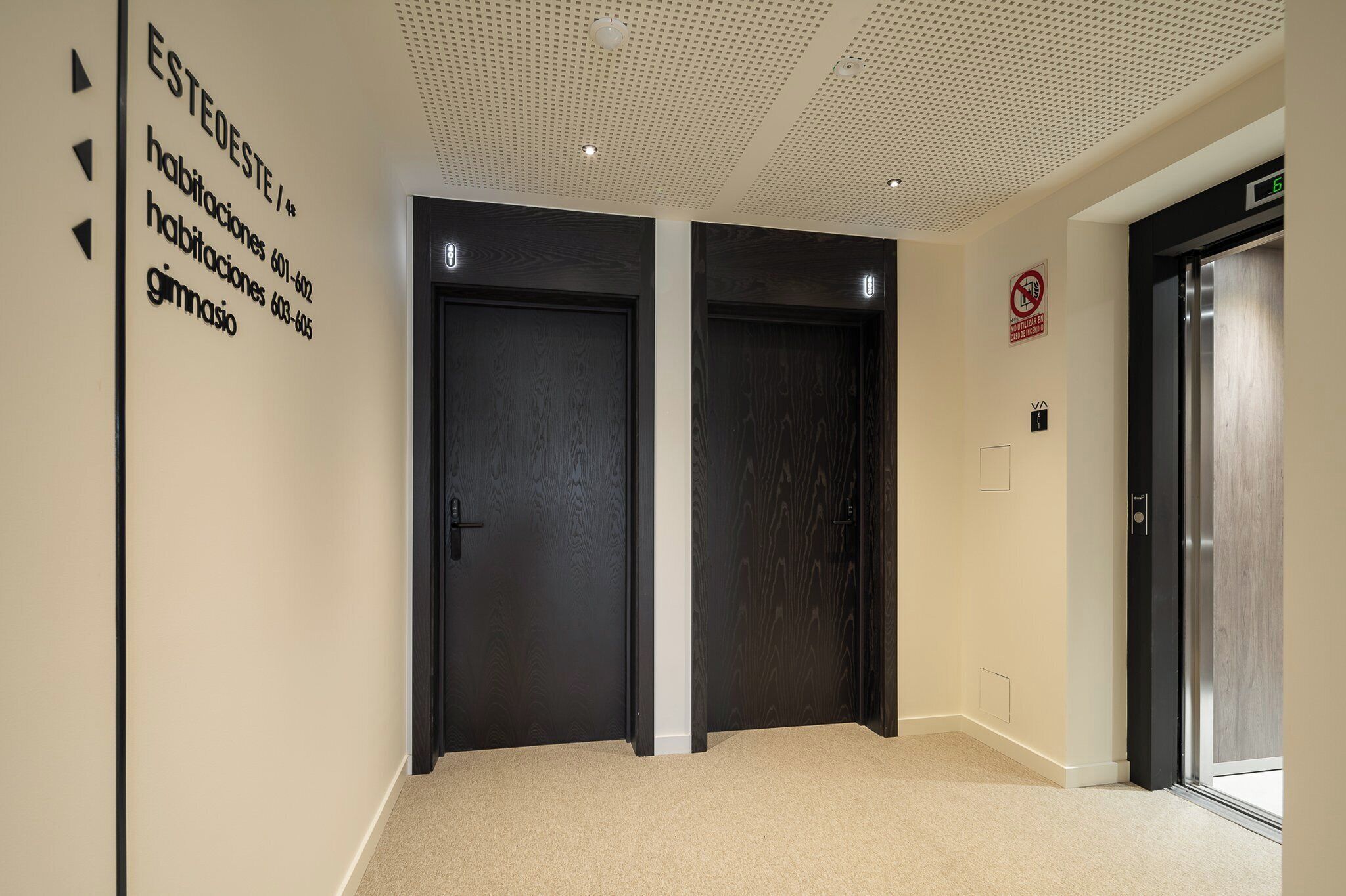 A hallway with two dark wooden doors, a wall with directional arrows and contact information, and an elevator with a digital display.