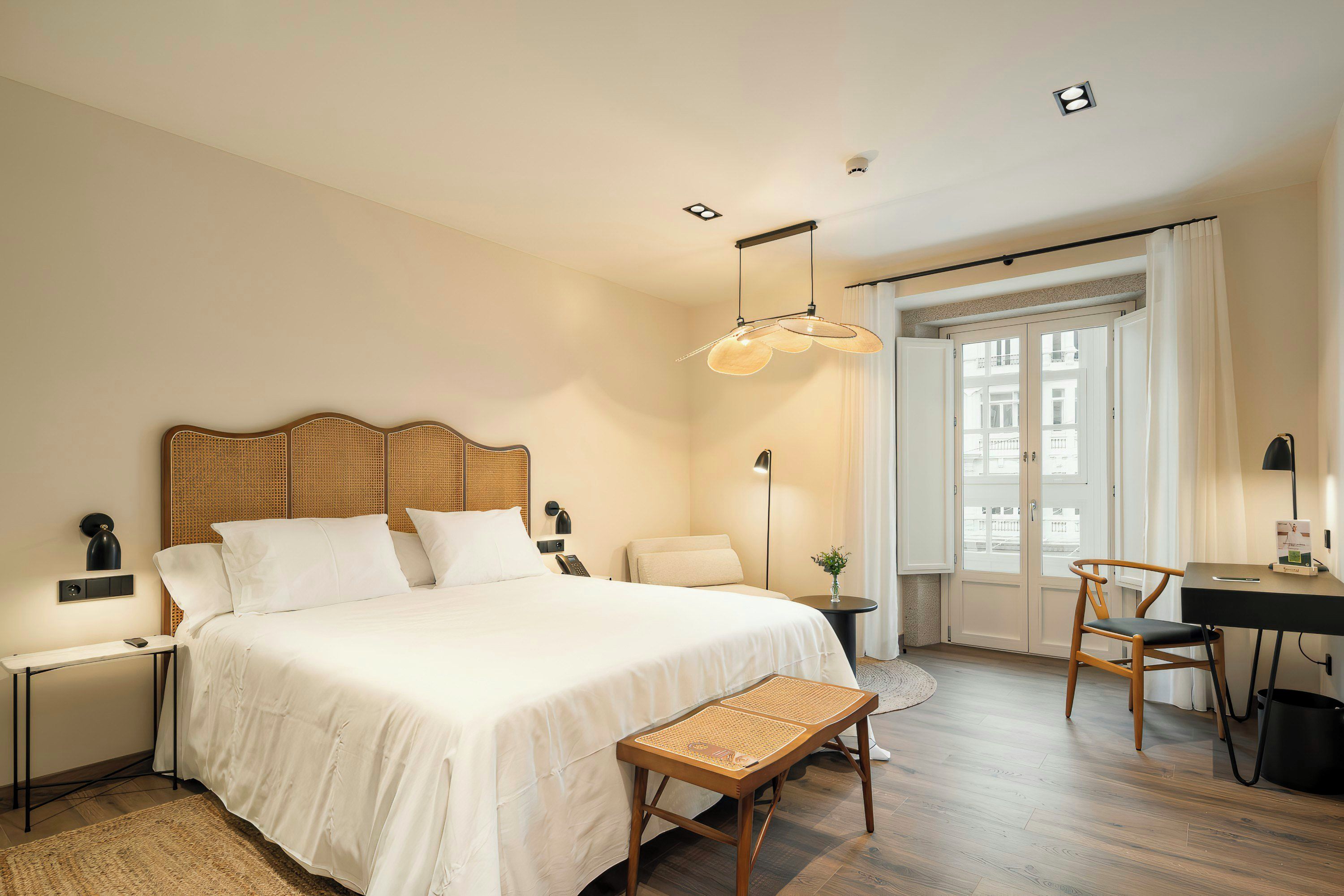 A hotel room with a large bed, a desk, a chair, a small table, and a window with curtains.