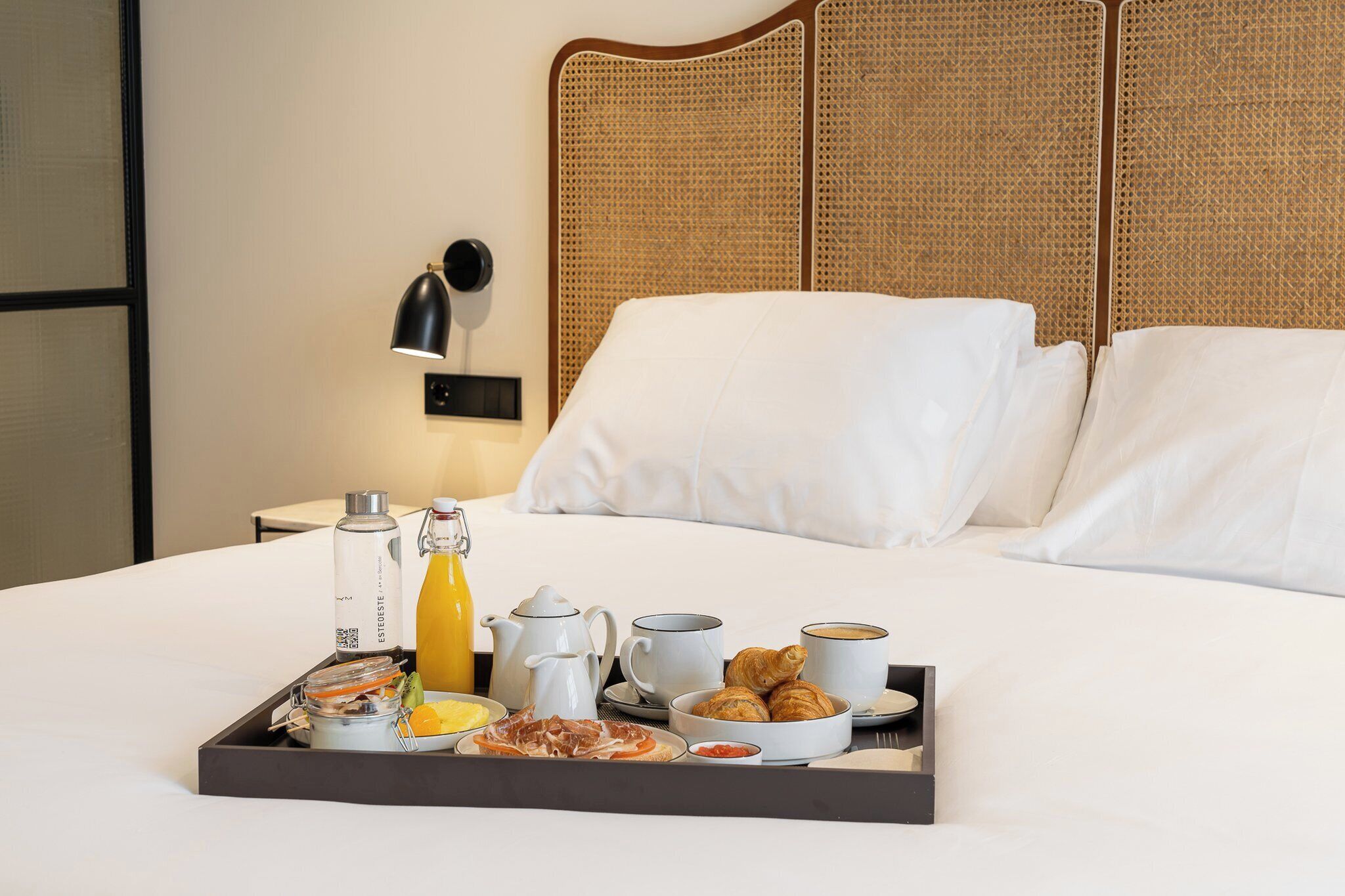 A neatly made bed with white linens, a tray with breakfast items including croissants, a bottle of orange juice, and a coffee maker.