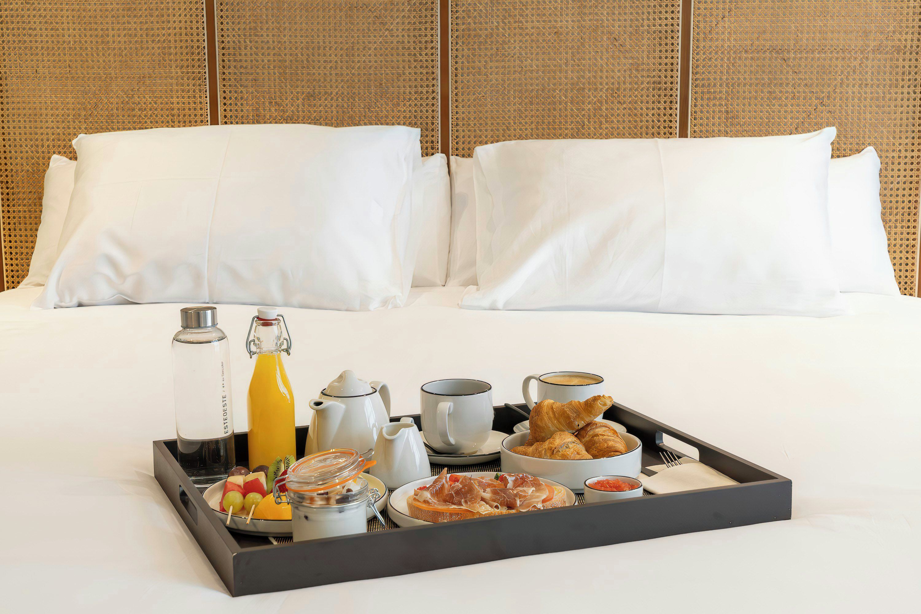 Un plateau de petit-déjeuner soigneusement préparé est posé sur un lit aux draps blancs, dont la tête de lit est ornée d’un motif tissé.