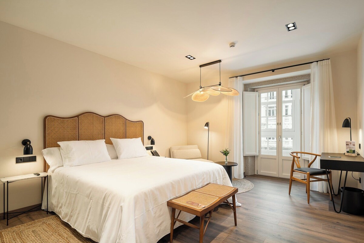 A hotel room with a large bed, a desk, a chair, a small table, and a bench.