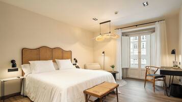 A hotel room with a large bed, a desk, a chair, a small table, and a bench.