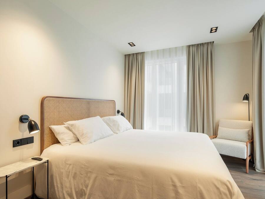 Una habitació d'hotel ben ordenada, amb un llit gran, una tauleta de nit, una cadira i una finestra amb cortines.