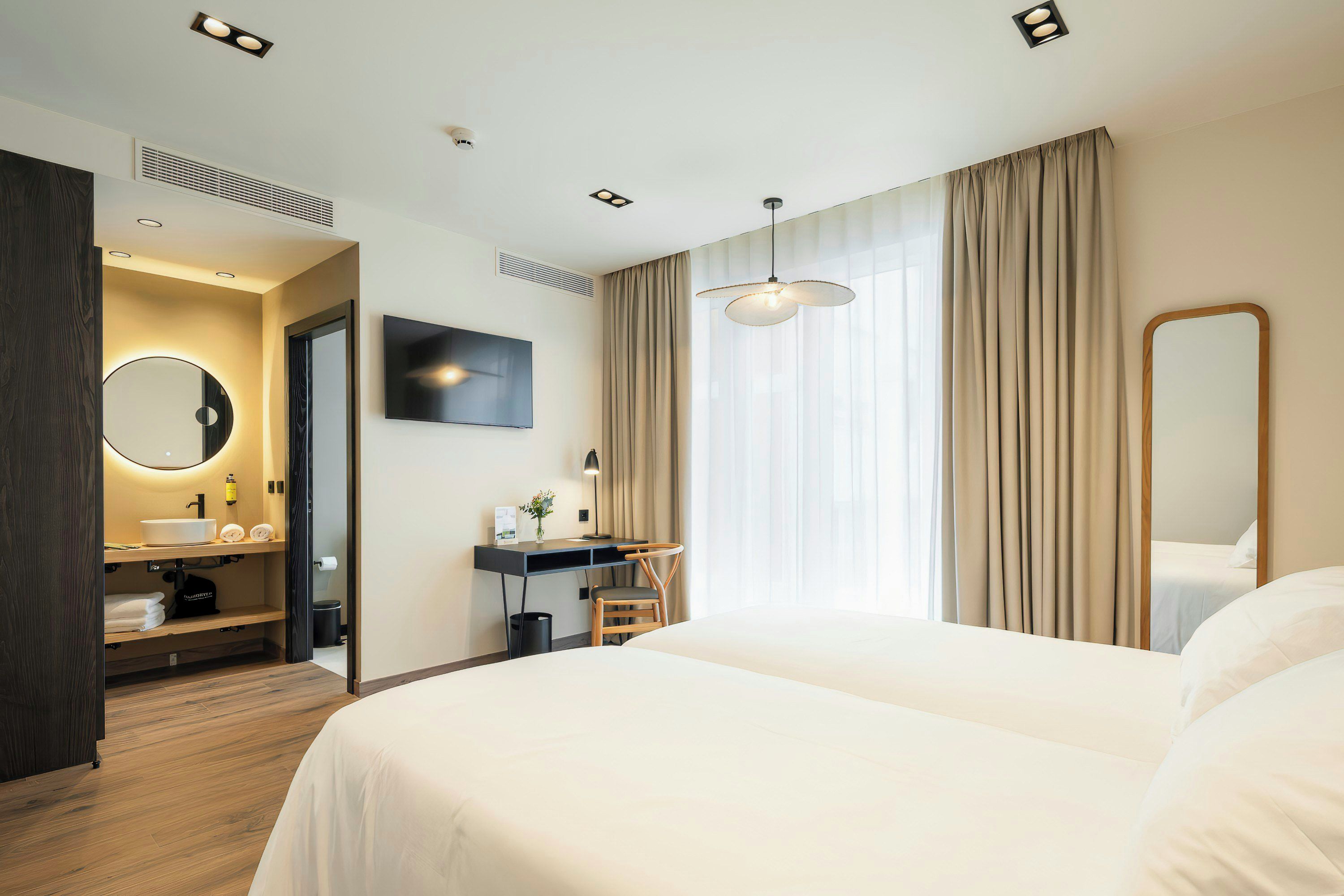A hotel room with a bed, a desk with a chair, a TV, a mirror, and a bathroom with a sink and toilet.