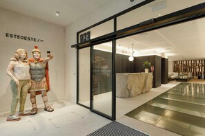 A modern interior with a reception desk, a seating area, and a glass wall displaying a sculpture. - Hotel Sercotel EsteOeste (Lugo)