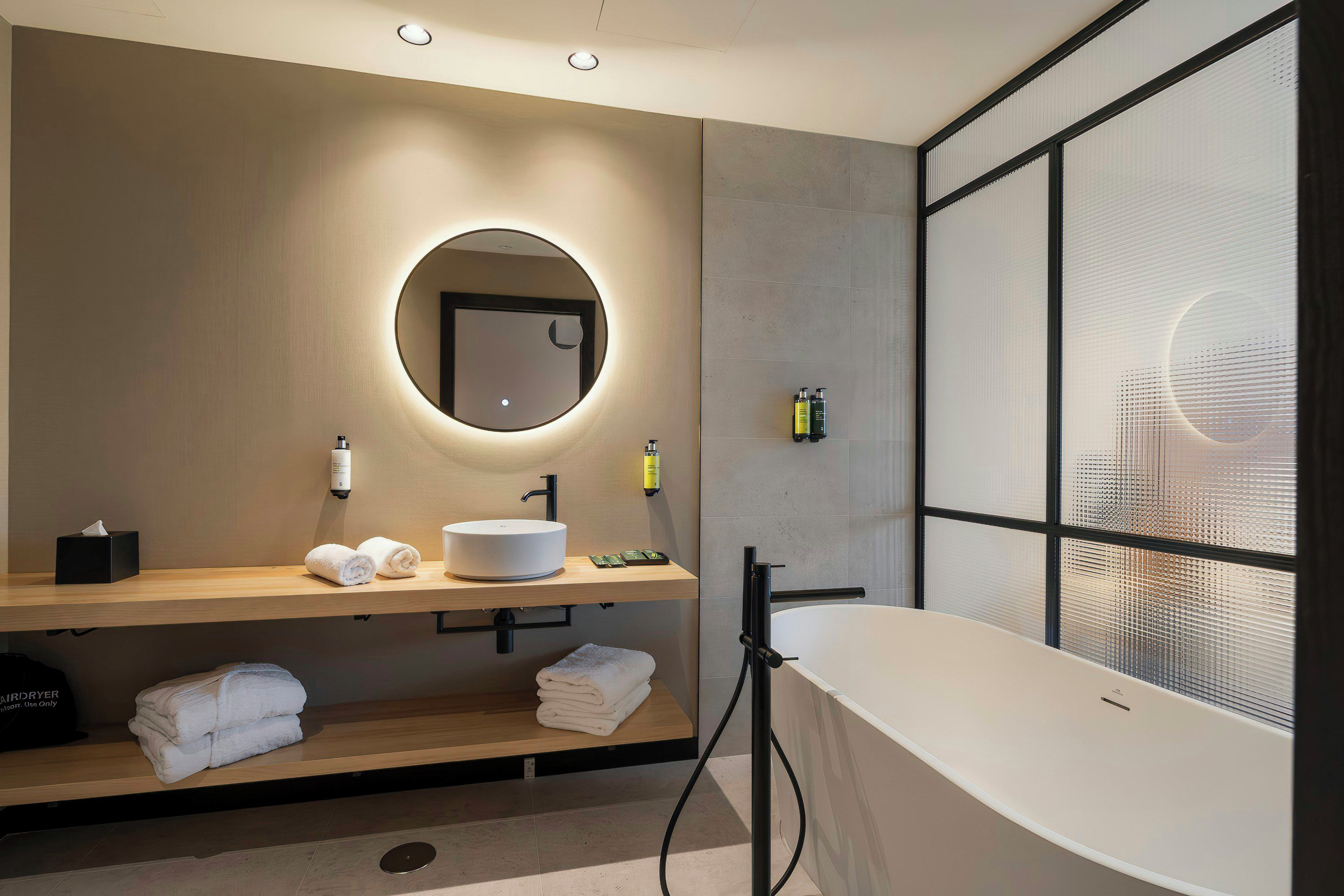 A modern bathroom with a round mirror, a white sink, a freestanding bathtub, and a window with a view.