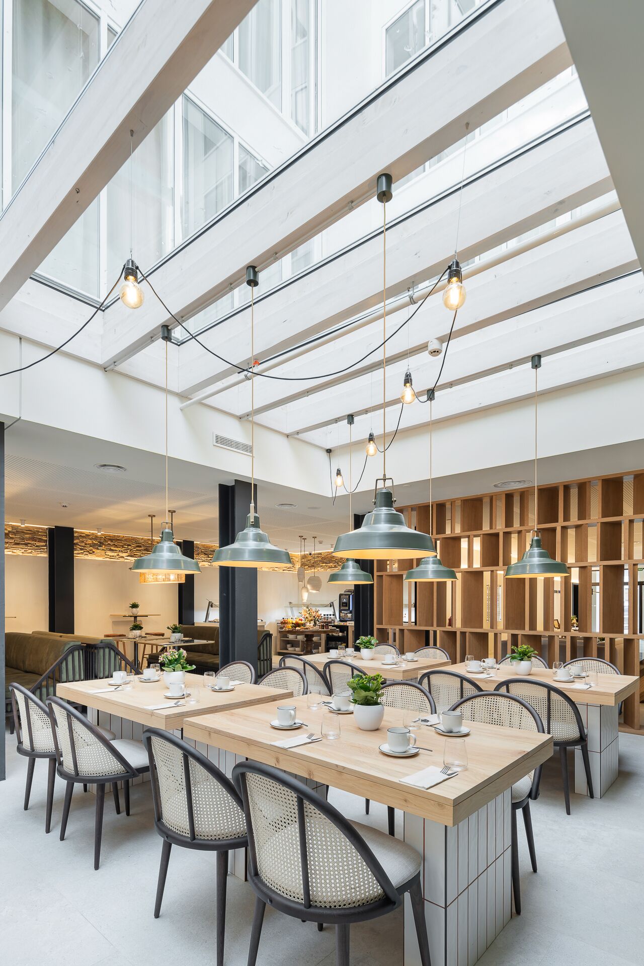 Uma sala de jantar moderna com pé direito alto, vigas aparentes e iluminação pendente. Mesas dispostas para vários convidados, e o espaço bem iluminado pela luz natural que entra por grandes janelas.