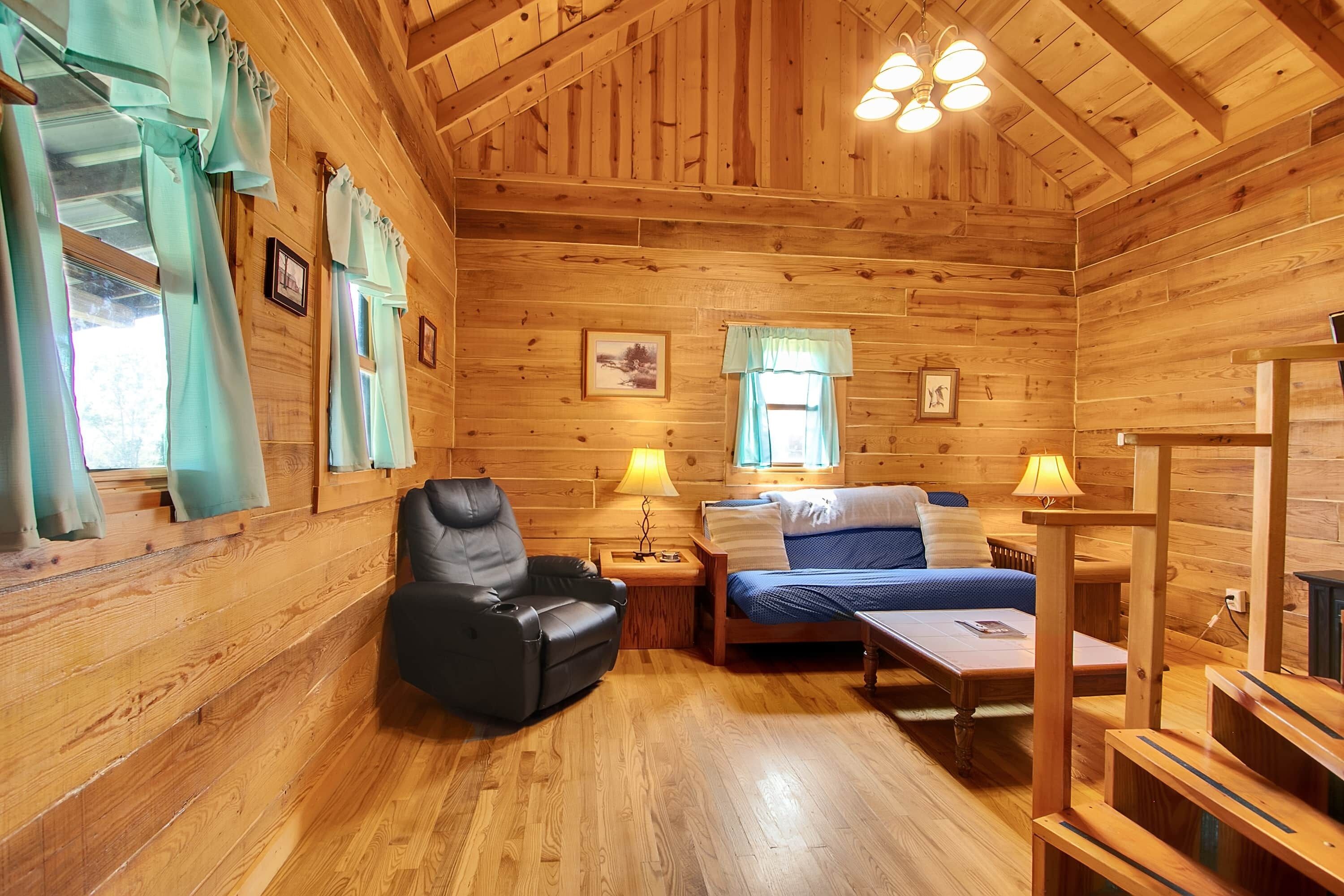 A wooden cabin interior with a bed, a chair, a table, and a lamp.