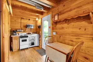 A compact kitchen with a stove, microwave, and oven, a dining table with a chair, and wooden walls.