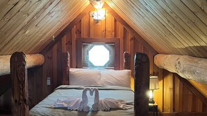 A cozy attic bedroom with a wooden ceiling, a large bed with white linens, and a window with blinds.