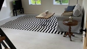 Un salón espacioso con un tapete a rayas negras y blancas, un sofá con cojines, una mesita redonda y dos ventanas con vistas a zonas verdes.