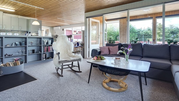 Un salon moderne avec un plafond en bois, un fauteuil blanc moelleux, un canapé gris et une table sur laquelle est posé un vase de fleurs.