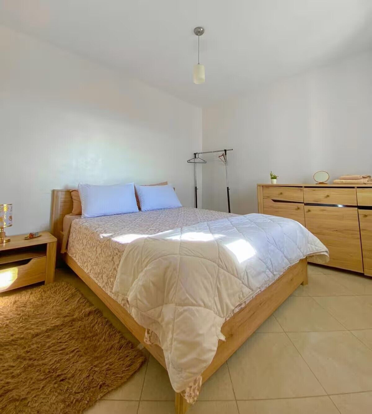 A bedroom with a wooden bed, a bedside table with a lamp, a dresser, and a hanging light fixture.