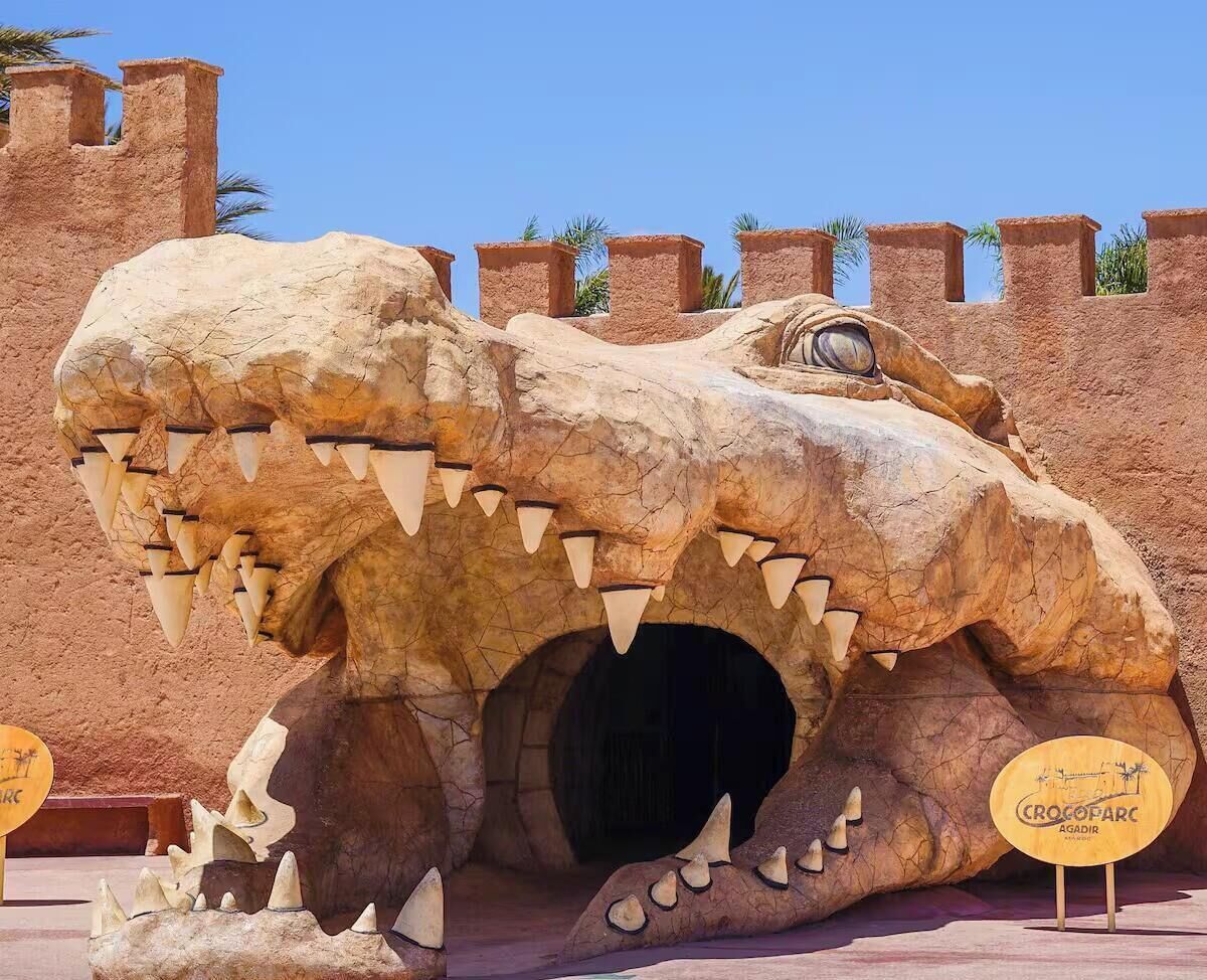 Een groot krokodillensculptuur met een grotopening, voor een muur met kantelen.