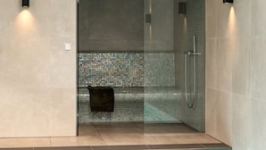 Une douche fermée par des parois de verre, avec un mur décoré d’une mosaïque de carreaux, une serviette accrochée à la porte, et une piscine située juste à côté de la douche.