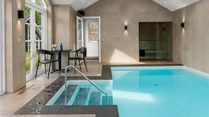 Un espace piscine intérieur moderne, offrant une vue dégagée sur le bassin, avec un coin salon doté de chaises et une porte donnant sur une autre pièce.