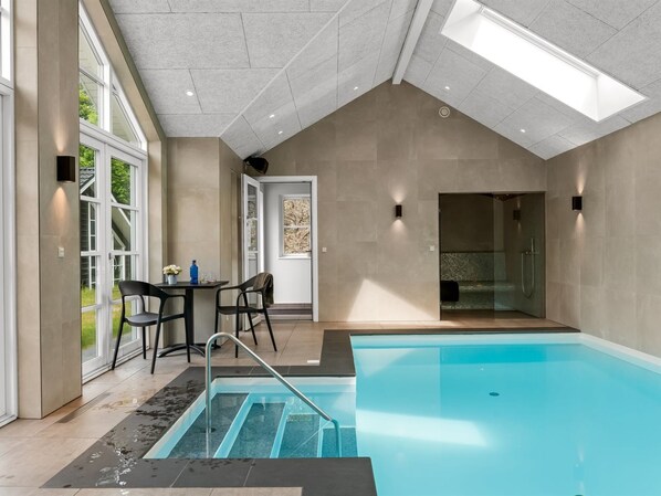 A modern indoor pool area with a clear view of the pool, a seating area with chairs, and a door leading to another room.
