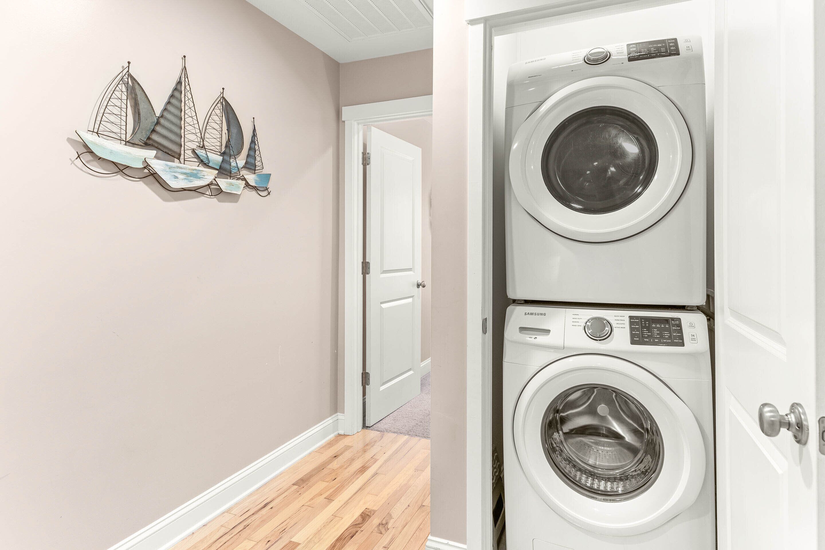 A laundry room with two stacked front-loading washing machines, a wooden floor, and a wall-mounted sailboat decoration.