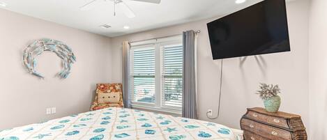 A bedroom with a bed, a dresser, a TV mounted on the wall, a ceiling fan, and a window with blinds.