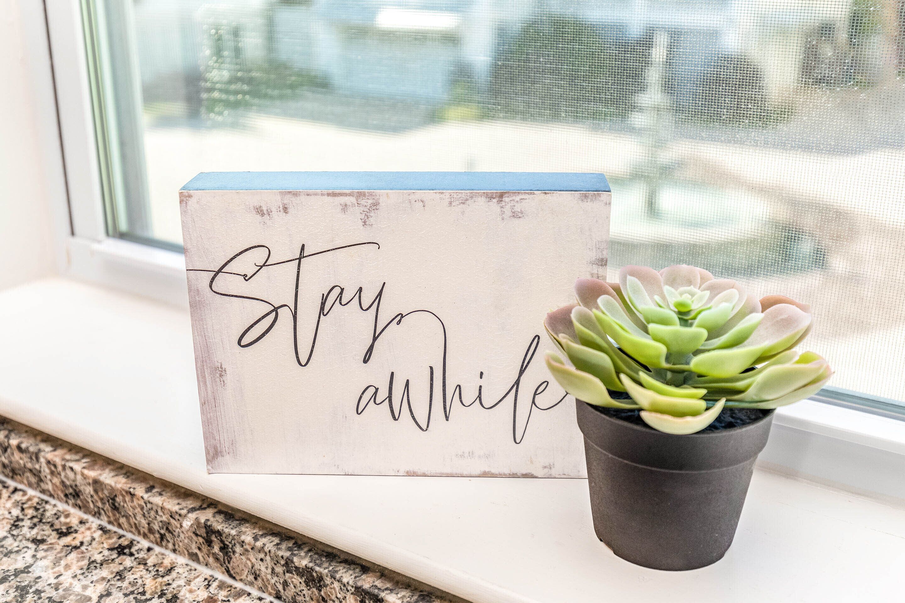 A sign with the text "Stay awhile" and a potted succulent plant on a windowsill.