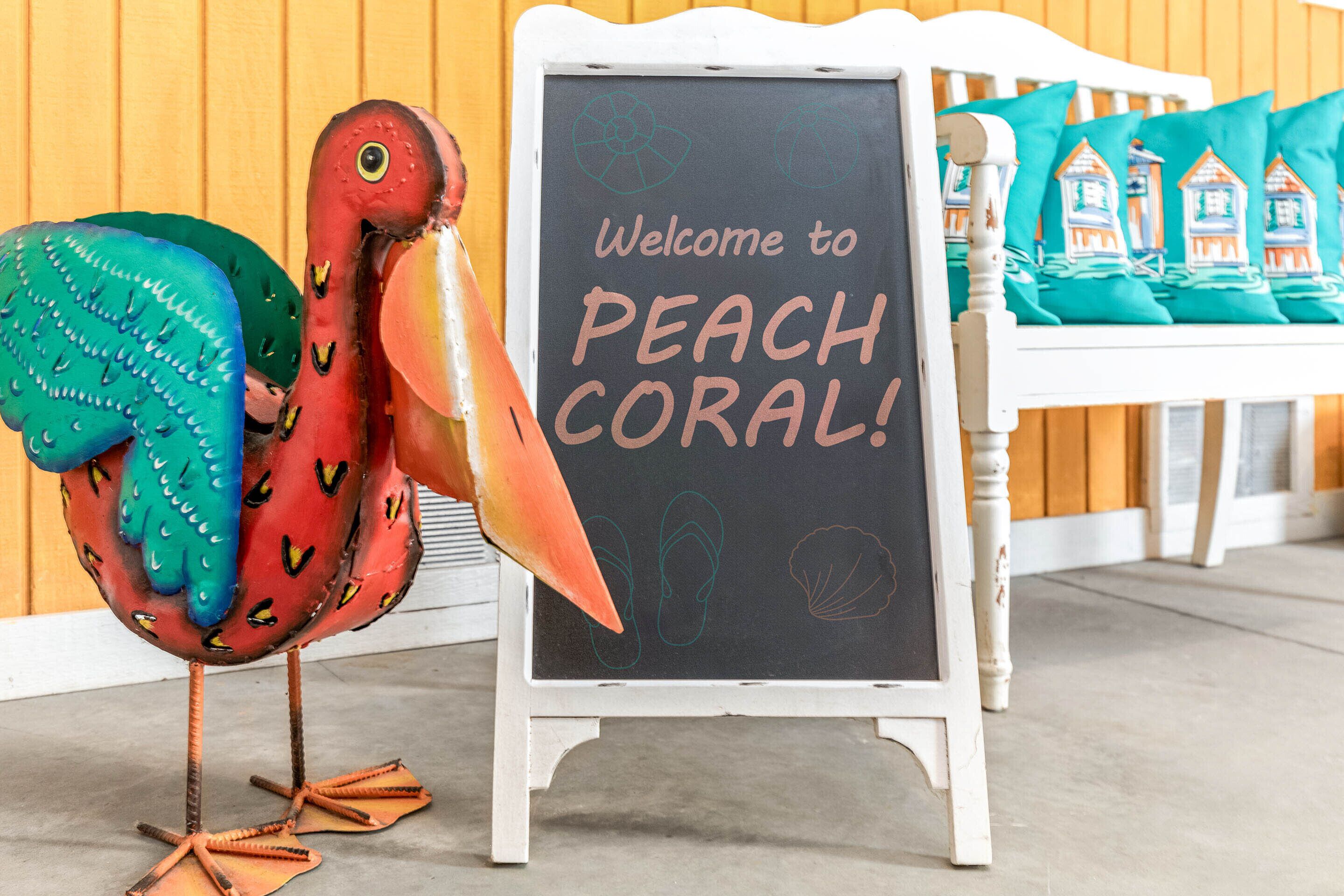 A chalkboard sign with "Welcome to PEACH CORAL!" written on it, a decorative bird, and teal pillows with house designs.