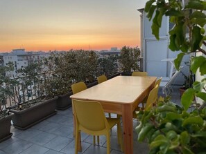 A rooftop terrace with a wooden table and yellow chairs, surrounded by potted plants and overlooking a cityscape at sunset. - Monopoli Roof - Penthouse with sea view and terrace (Monopoli)