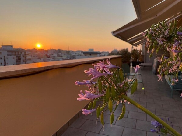 A rooftop terrace with a view of the sunset over a cityscape, featuring a water body and flowering plants. - Monopoli Roof - Penthouse with sea view and terrace (Monopoli)