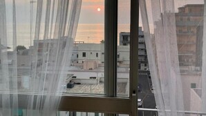 A view through a window with sheer curtains, featuring a sunset over a cityscape with buildings.