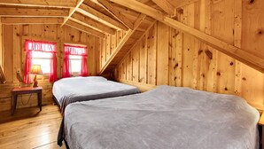 Uma cabana de madeira com duas camas, uma mesa pequena e uma janela com cortinas vermelhas.