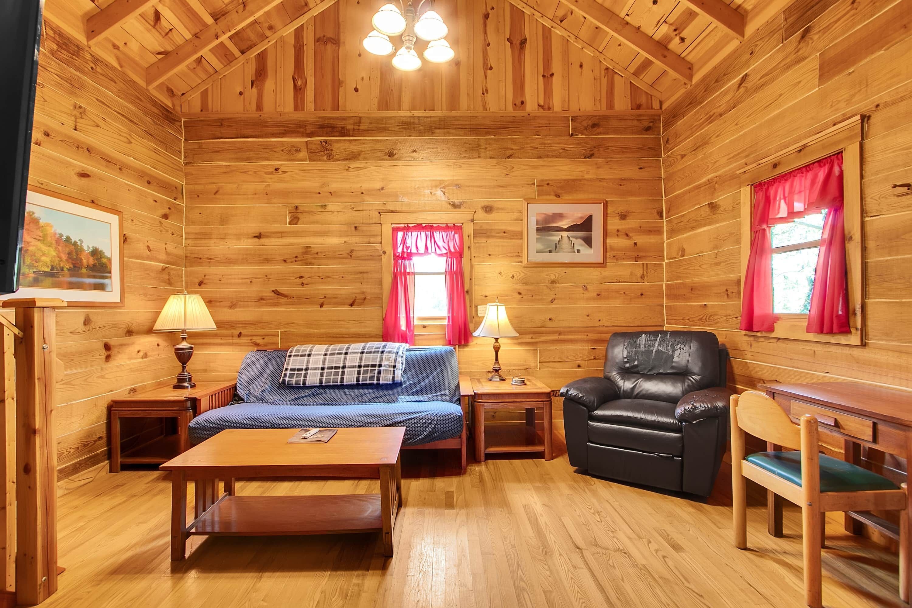 Interior de una cabaña de madera con sofá, sillón, mesa y cama.