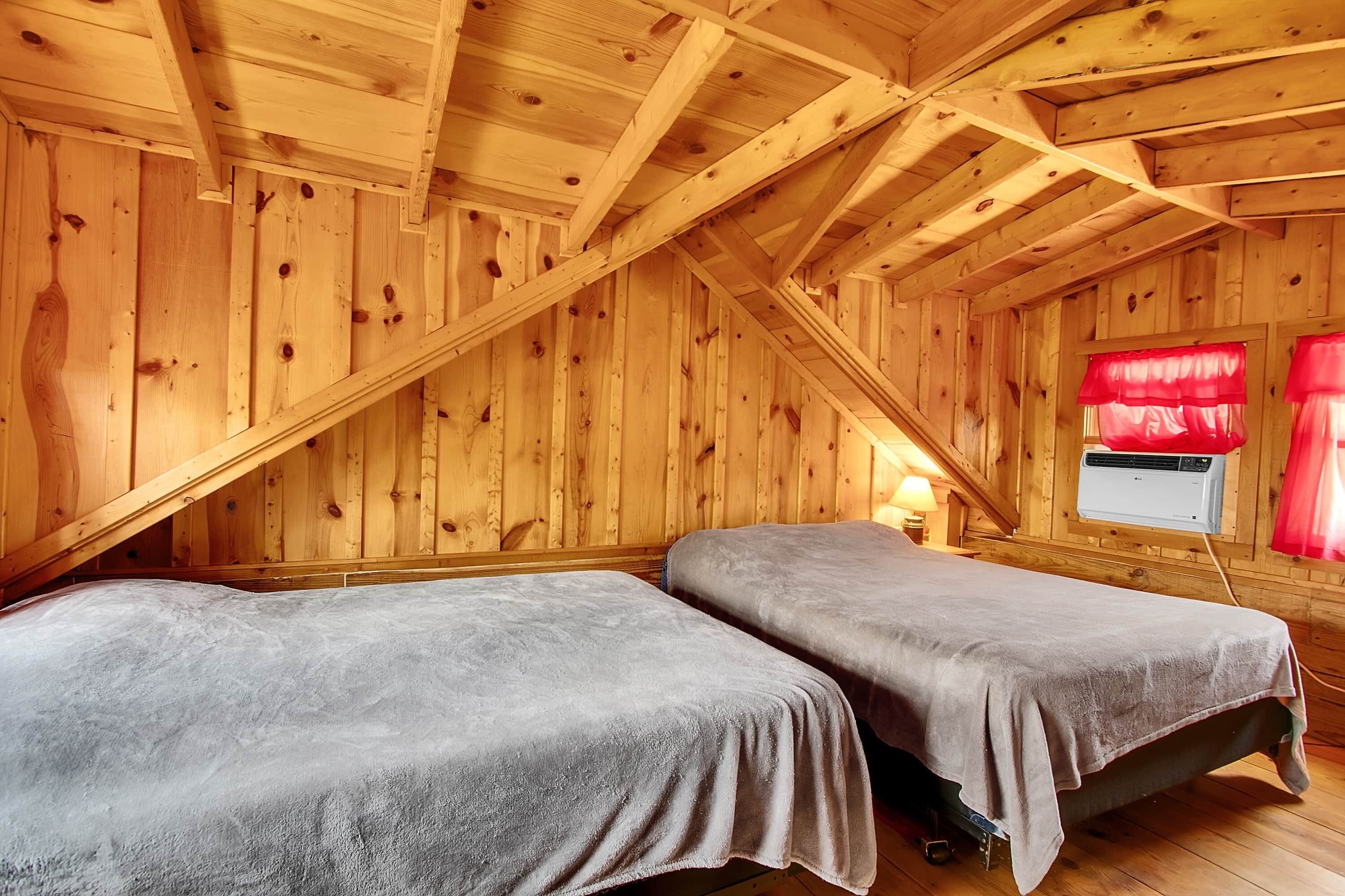 Une pièce aux murs en bois et au plafond en pente, comprenant deux lits avec une literie grise, une fenêtre avec des rideaux rouges et un appareil de climatisation.