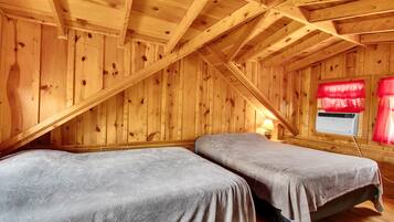 A room with wooden walls and a sloped ceiling, featuring two beds with grey bedding, a window with red curtains, and an air conditioning unit.