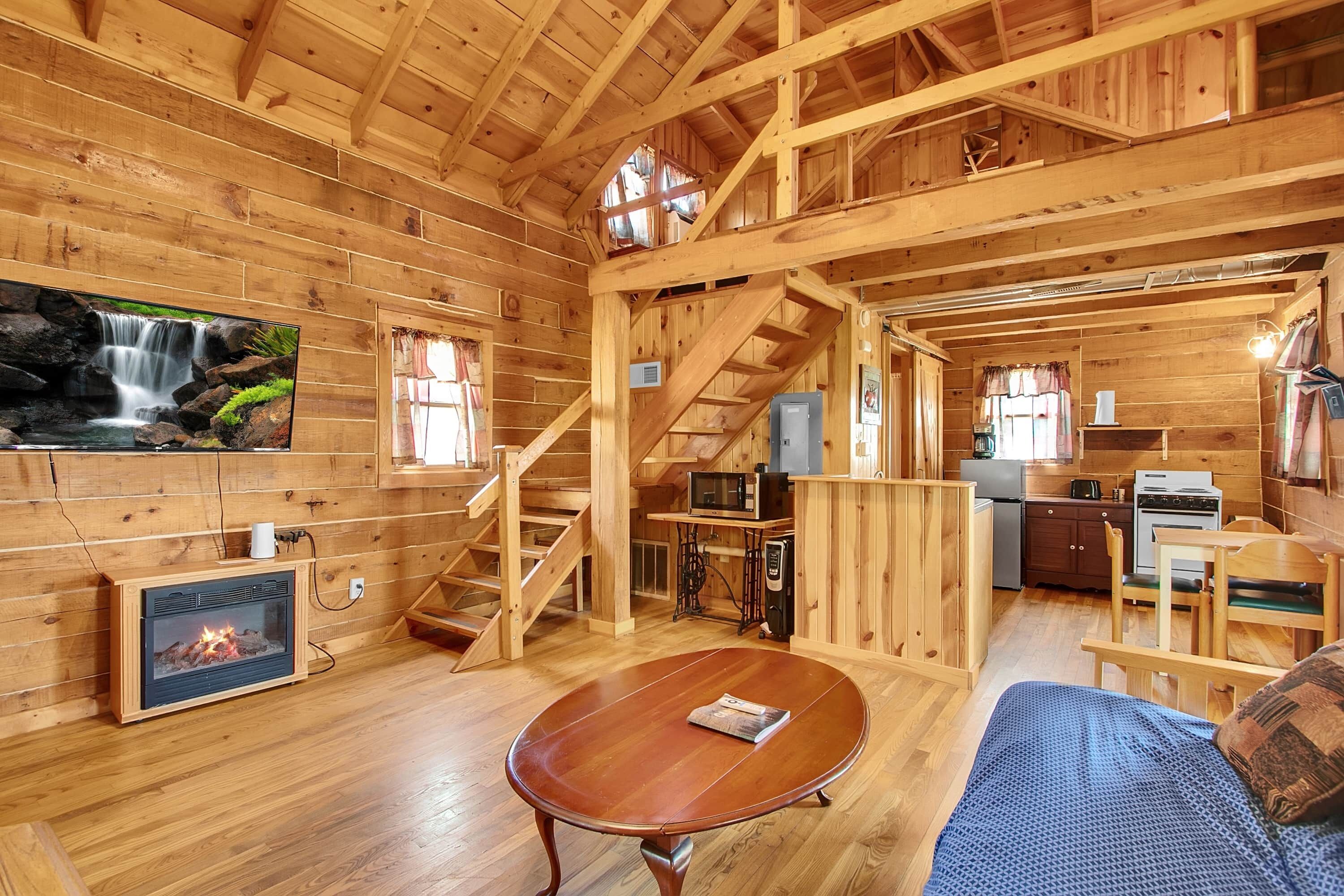 A wooden cabin interior with a fireplace, a dining area, a kitchen, and a staircase.