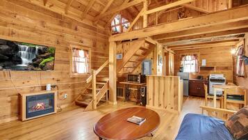 A wooden cabin interior with a fireplace, a dining area, a kitchen, and a staircase.