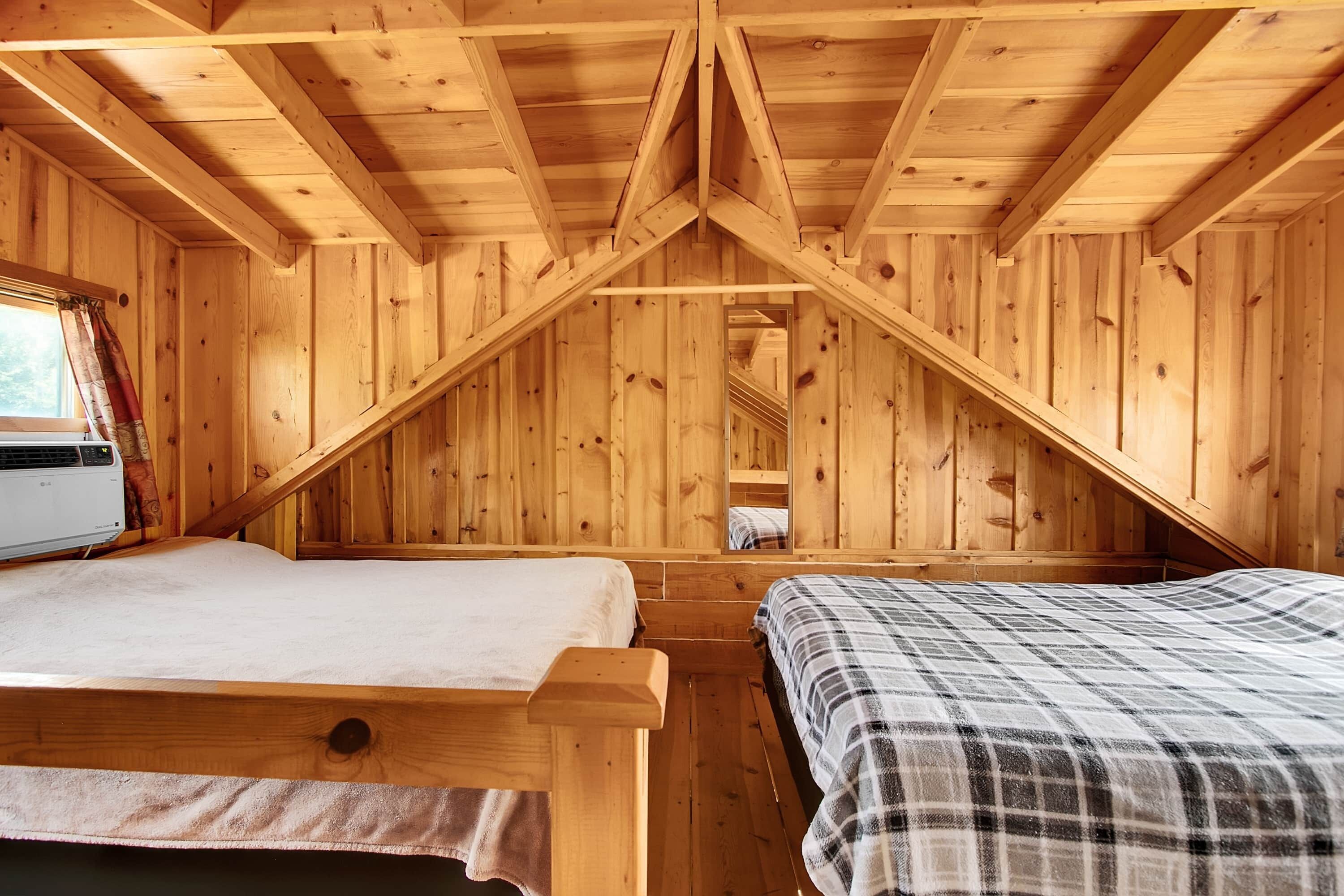 A wooden cabin room with a sloped ceiling, two beds, a window with curtains, and a visible air conditioning unit.