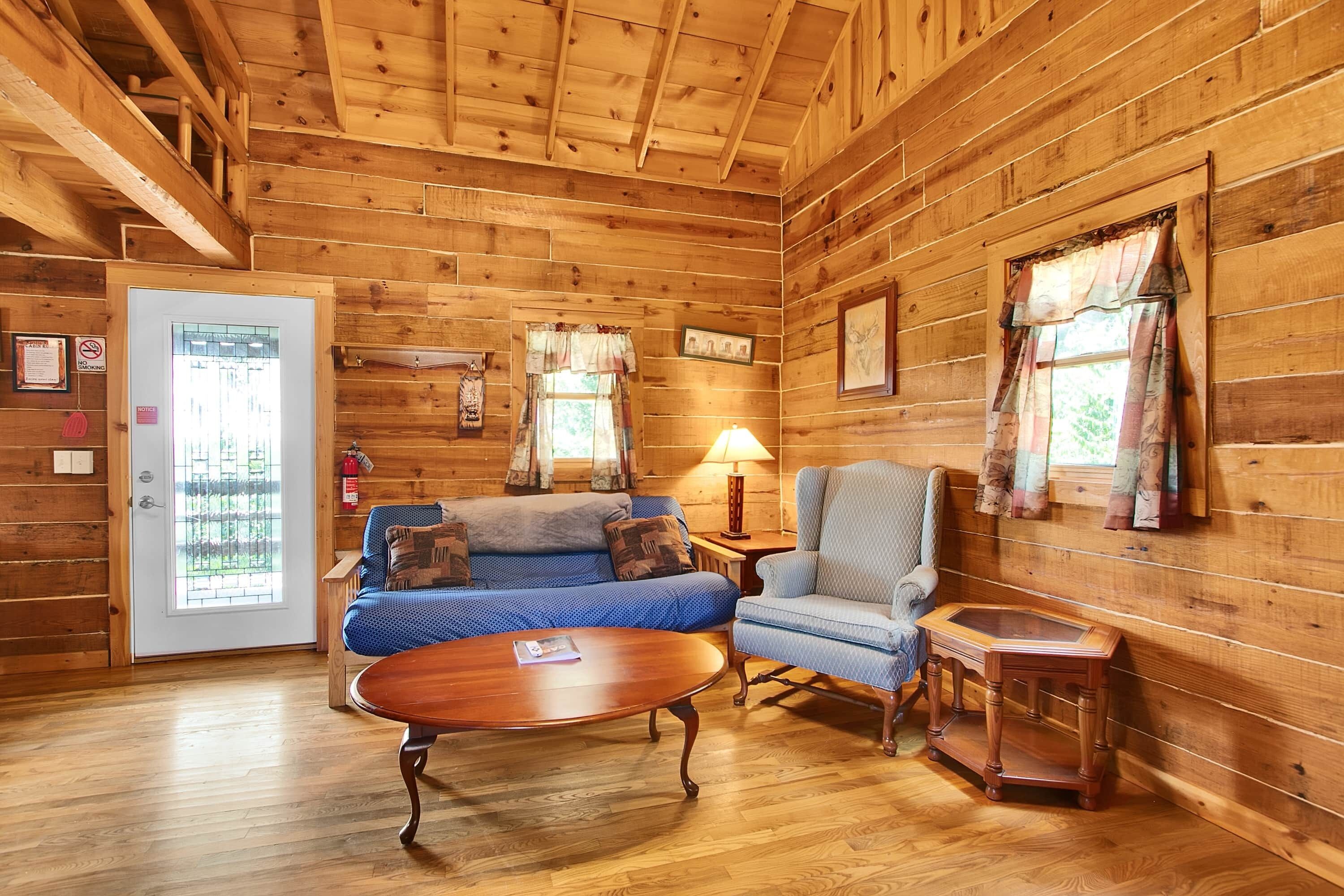 A log cabin interior with a wooden floor, a sofa, a chair, a small table, and a door with a window.
