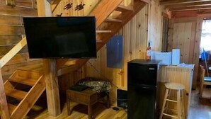 En stuga med träpanel på insidan, en trappa, en tv som hänger på väggen, ett kylskåp och en liten pall.