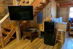 Ein Holzhütten-Innenraum mit Treppe, einem an der Wand montierten Fernseher, einem Kühlschrank und einem kleinen Hocker.