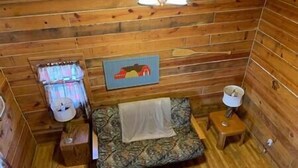 En stuga med träpanel på insidan, inredd med en säng, ett litet bord, en lampa och en tavla på väggen.