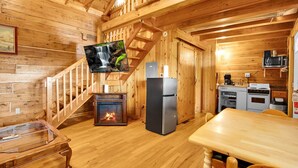 Interior de uma cabana de madeira com lareira, televisão na parede, área de cozinha com eletrodomésticos e escada de madeira.