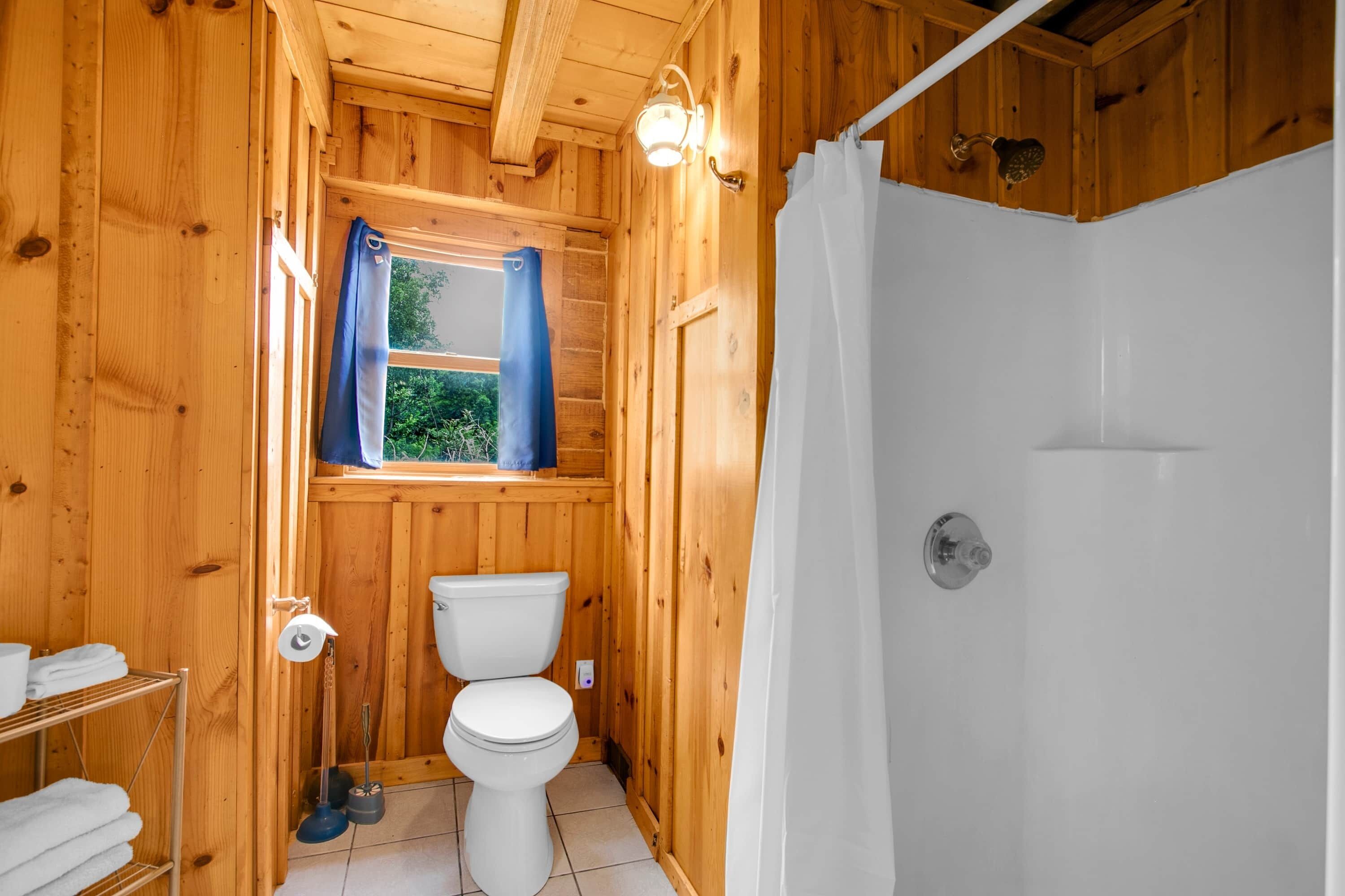 Ein Badezimmer einer Holzhütte mit Toilette, Dusche und einem Fenster mit Vorhängen.