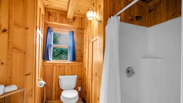 Ein Badezimmer einer Holzhütte mit Toilette, Dusche und einem Fenster mit Vorhängen.