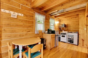 Interior de una cabaña de madera con una mesa de comedor, sillas, y una zona de cocina que incluye estufa, microondas y refrigerador.