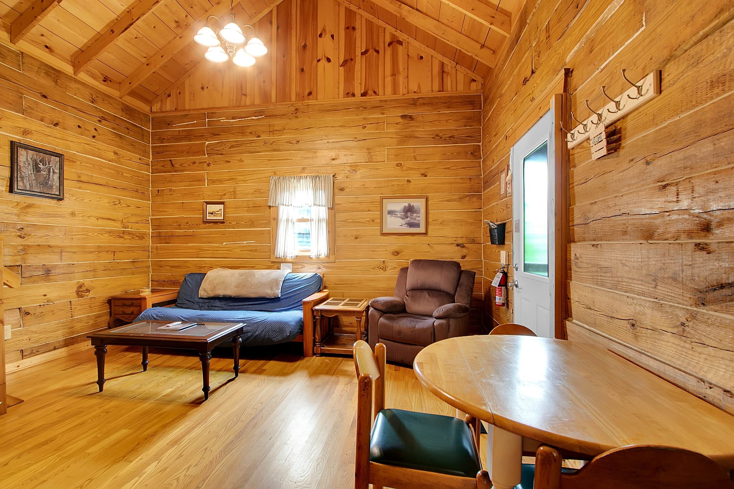 Interior d'una cabana de fusta amb un llit, una cadira, una taula i una finestra amb cortines.