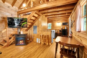 A wooden cabin interior with a fireplace, a TV displaying a waterfall, a kitchen area, and a staircase.