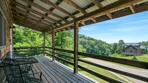 Um deck de madeira com mesa e cadeiras de metal, com vista para uma paisagem verde e uma estrada.