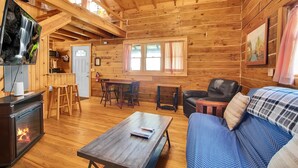 Interior de uma cabana de madeira com lareira, sofá, área de jantar e televisão.