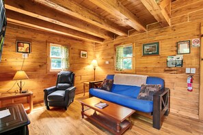 A wooden cabin interior with a sofa, chair, table, lamp, framed pictures, and a fire extinguisher.