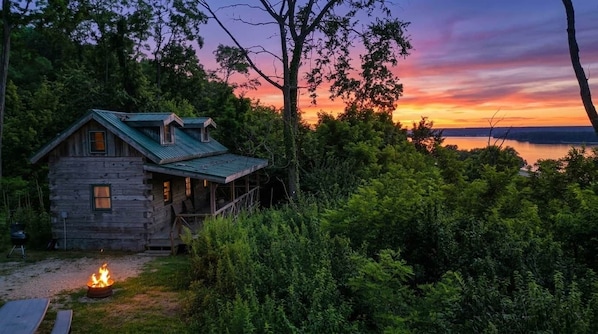 A rustic cabin with a fire pit, surrounded by dense greenery and overlooking a body of water.